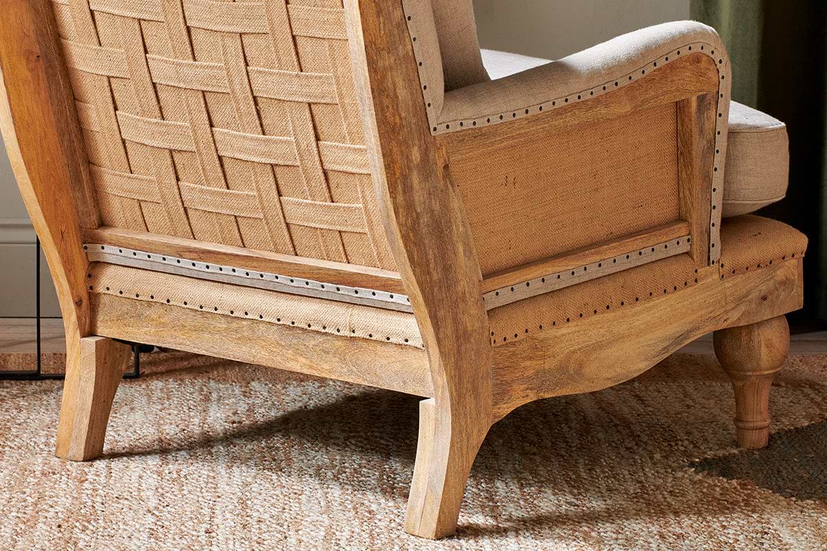 The Abe Deconstructed Linen Armchair - Stone by nkuku features a sturdy mango wood frame, curved legs, and linen upholstery with visible natural wood grain. It sits on a textured beige rug, showcasing a distinctive basket-weave pattern on the back.