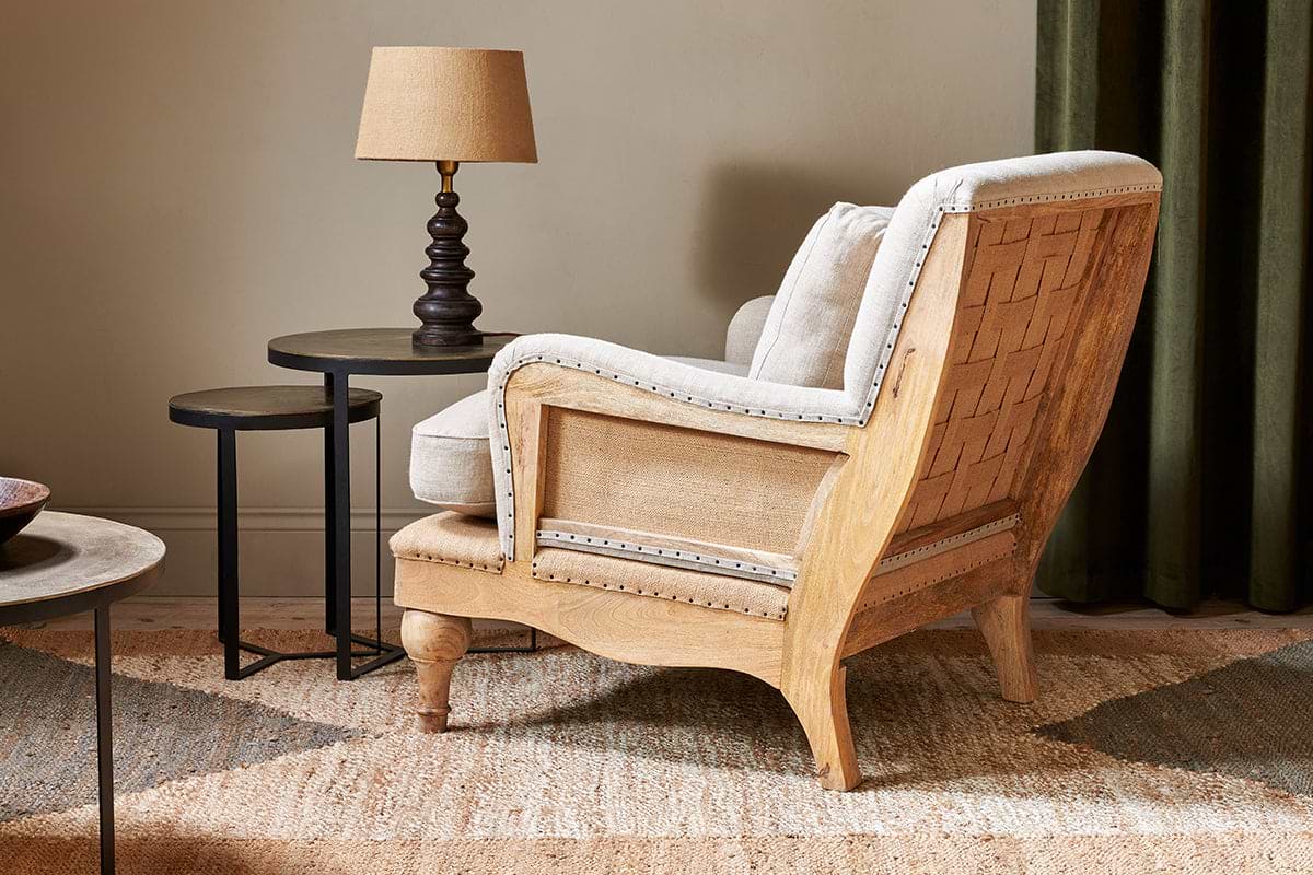 The nkuku Abe Deconstructed Linen Armchair in Stone rests on a woven rug next to two round black nesting tables and a brown lamp. The room features neutral walls and dark green curtains.