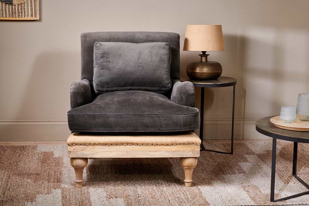 The nkuku Abe Deconstructed Velvet Armchair in moss, featuring a matching back cushion, pairs with a light wood ottoman and round side table topped by a textured brown lamp, all resting on a patterned beige rug against a neutral wall.