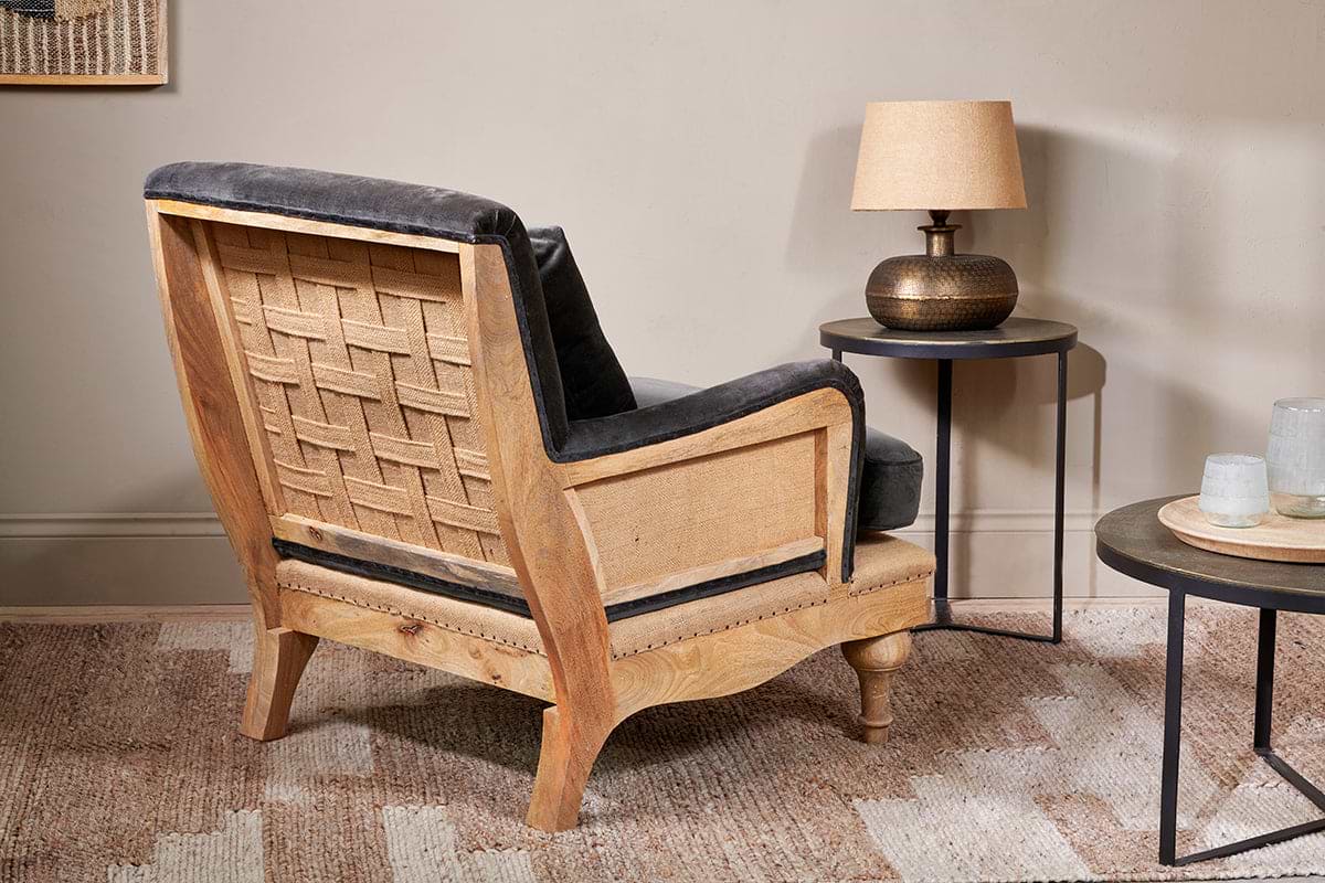 The Abe Deconstructed Velvet Armchair in moss by nkuku faces a round side table with a bronze lamp and beige shade. A textured rug covers the floor, and another table with glass items is partially visible.
