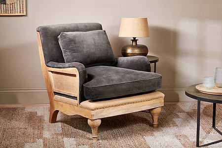 The Abe Deconstructed Velvet Armchair by nkuku in moss sits on a patterned rug with dark gray cushions. A round side table with a lamp and glassware stands nearby, while a light beige wall enhances the cozy, inviting atmosphere.
