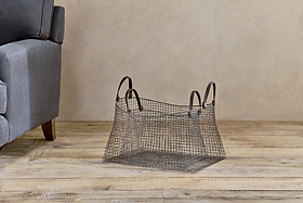 The Adah Iron Baskets - Brown (Set of 2) by nkuku, featuring leather handles and a geometric design, sit beside a gray sofa on a light wood floor, complementing the neutral, minimalist decor against the beige wall.