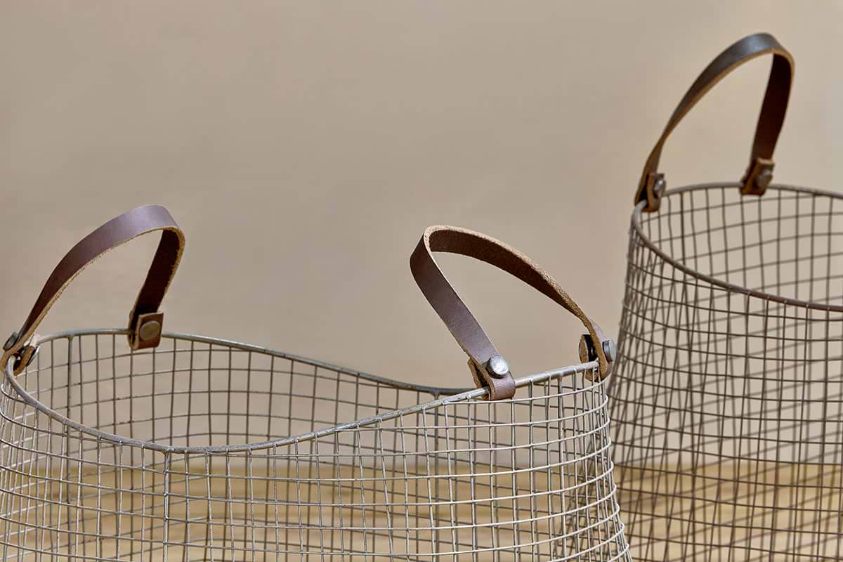 The Adah Iron Baskets - Brown (Set of 2) by nkuku sit on a light wooden surface, showcasing their open grid design and sturdy brown leather handles—perfect stackable storage with only the tops and handles visible.