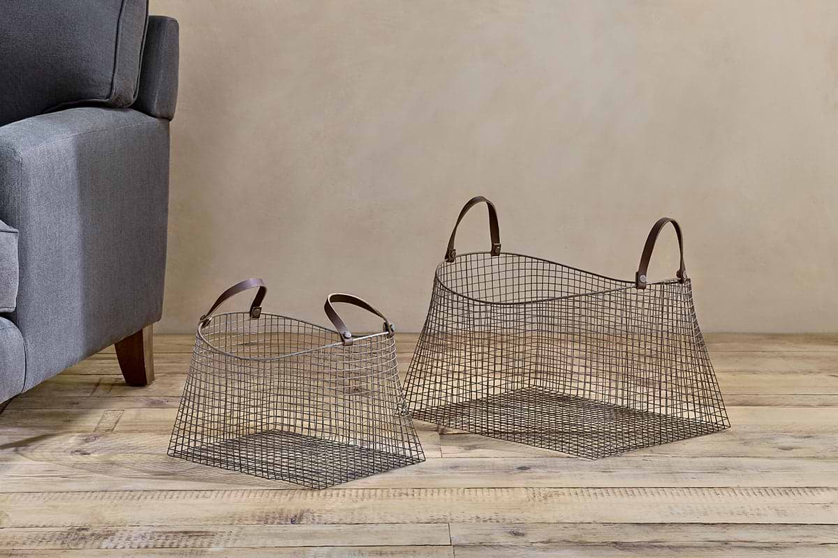The Adah Iron Baskets - Brown (Set of 2) by nkuku, featuring leather handles and a grid design, sit on a wooden floor beside a grey armchair. These stackable baskets come in different sizes.