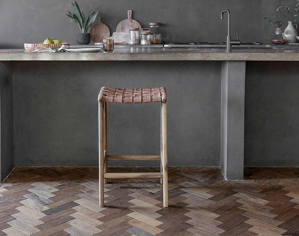 The Adembi Woven Leather Counter Stool by nkuku, with its wooden frame, stands on a herringbone floor near a gray concrete kitchen counter topped with dishes, glassware, potted plants, accessories, and a modern faucet in the background.