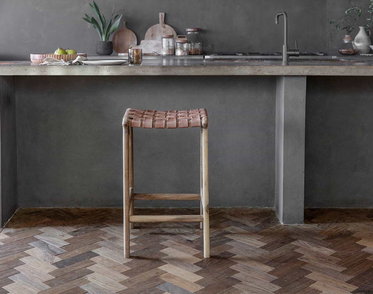 The Adembi Woven Leather Counter Stool by nkuku, with its wooden frame, stands on a herringbone floor near a gray concrete kitchen counter topped with dishes, glassware, potted plants, accessories, and a modern faucet in the background.