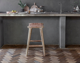The Adembi Woven Leather Counter Stool by nkuku sits beside a concrete countertop in a modern kitchen with gray walls, chevron wood flooring, and minimalist decor featuring plants, fruit, and cutting boards.