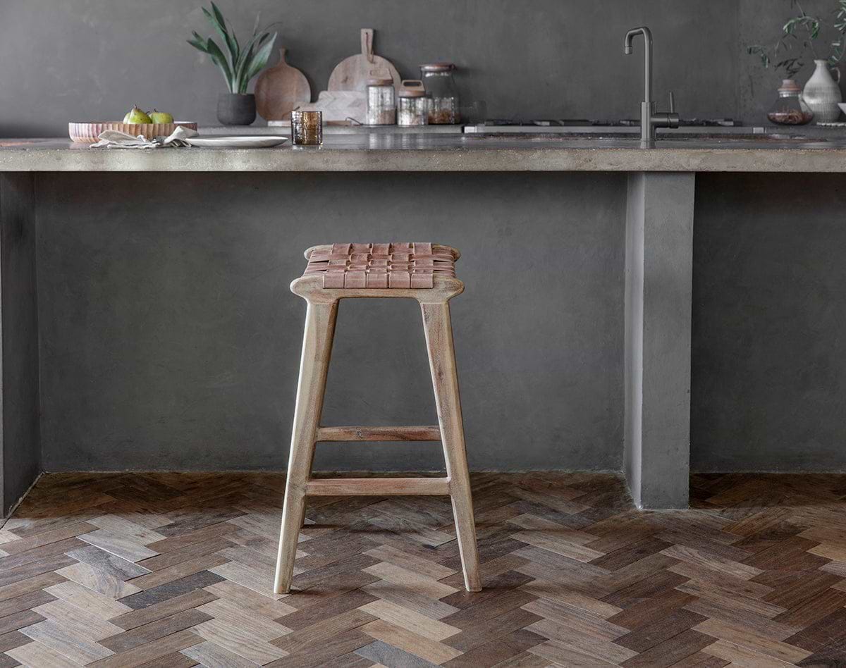 The Adembi Woven Leather Counter Stool by nkuku sits beside a concrete countertop in a modern kitchen with gray walls, chevron wood flooring, and minimalist decor featuring plants, fruit, and cutting boards.