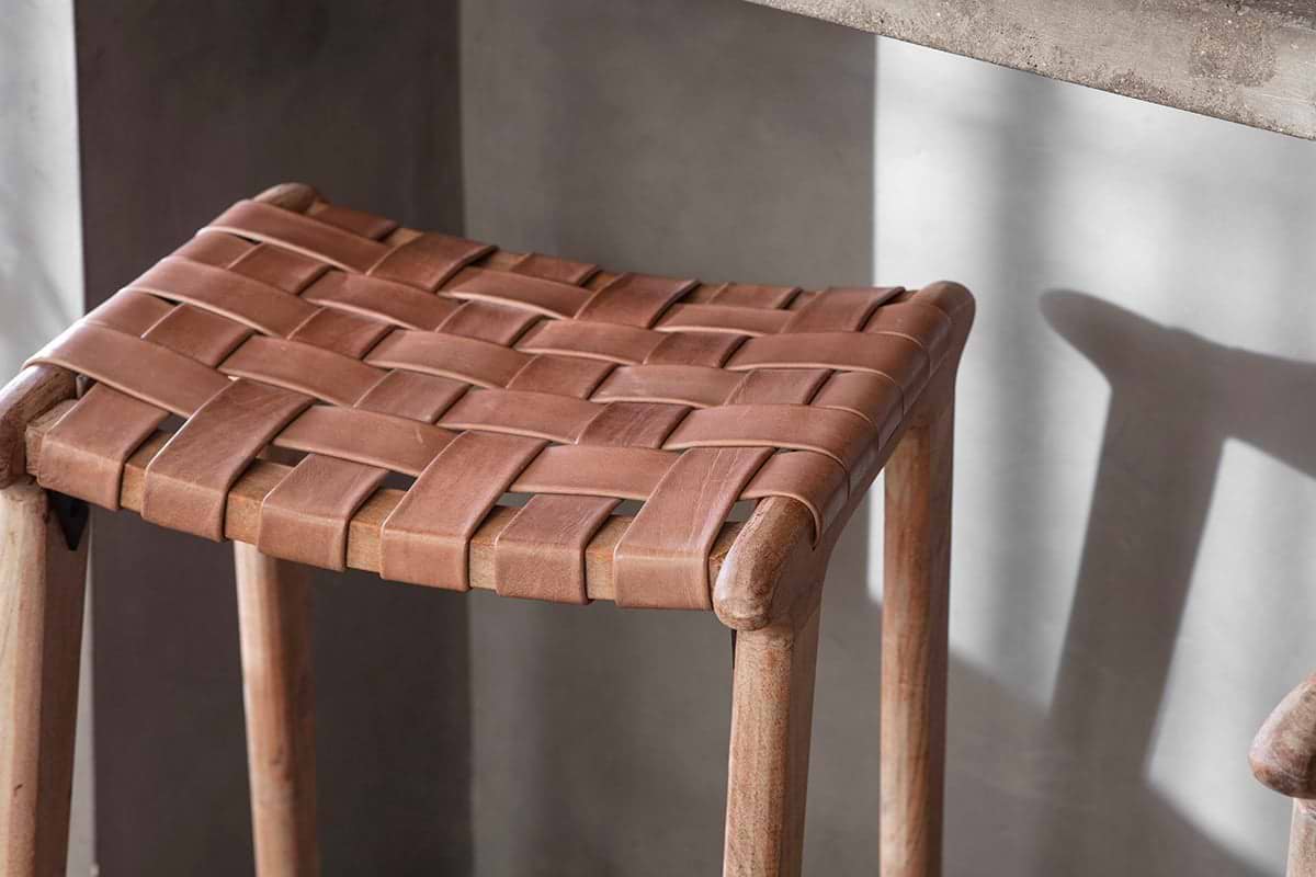 A close-up of the nkuku Adembi Woven Leather Counter Stool shows its tan, tightly woven leather seat in a checkered pattern atop rounded light acacia wood legs, with soft light casting gentle shadows on the neutral gray wall behind.