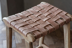 A close-up of the nkuku Adembi Woven Leather Counter Stool, showcasing wide tan leather straps in a crisscross pattern and a natural-finish wood frame, set on an elegant herringbone hardwood floor.