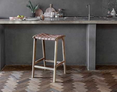 A rustic kitchen showcases nkuku's Adembi Woven Leather Counter Stool with a wooden frame. A concrete countertop, sink, kitchen items, and a plant in a vase complete the cozy space, while a herringbone wood floor adds warmth.