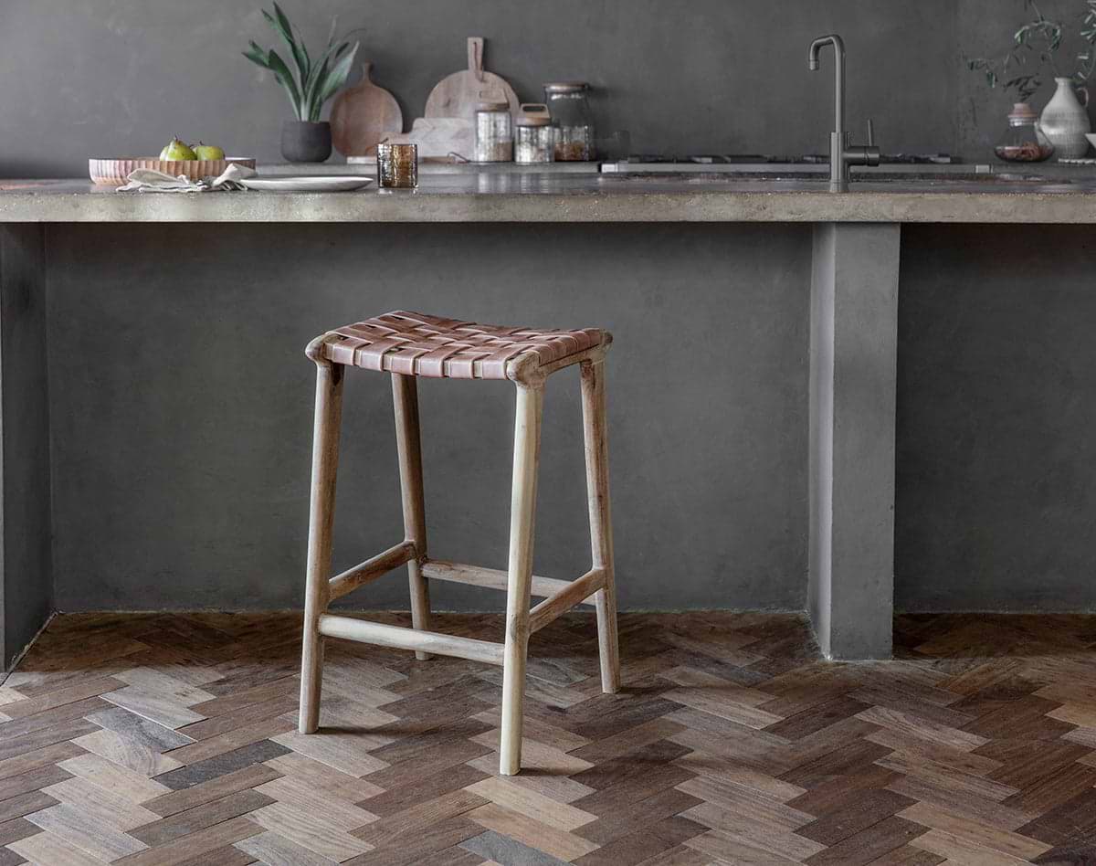 A rustic kitchen showcases nkuku's Adembi Woven Leather Counter Stool with a wooden frame. A concrete countertop, sink, kitchen items, and a plant in a vase complete the cozy space, while a herringbone wood floor adds warmth.