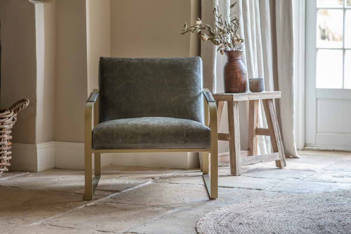 The nkuku Adra Leather & Brass Occasional Chair in moss green sits on a stone floor. To the right is a wooden side table with a vase of dried flowers and a small dark pot.