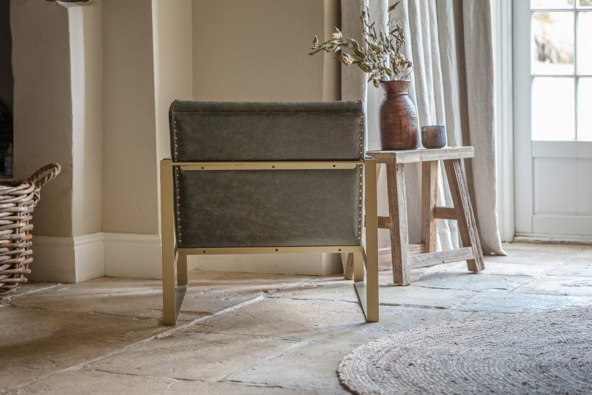 A rustic room with an nkuku Adra Leather & Brass Occasional Chair in moss green facing away, antique brass stud accents, stone tile floor, woven basket, wood side table with brown vases and dried branches, a round textured rug, and sunlight through light curtains.