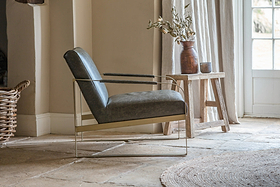 The nkuku Adra Leather & Brass Occasional Chair in Moss Green, featuring slender brass arms and legs, sits on a stone floor by a rustic wood table adorned with a brown vase of dried flowers and a ceramic jug. Sunlight filters through glass panes behind.