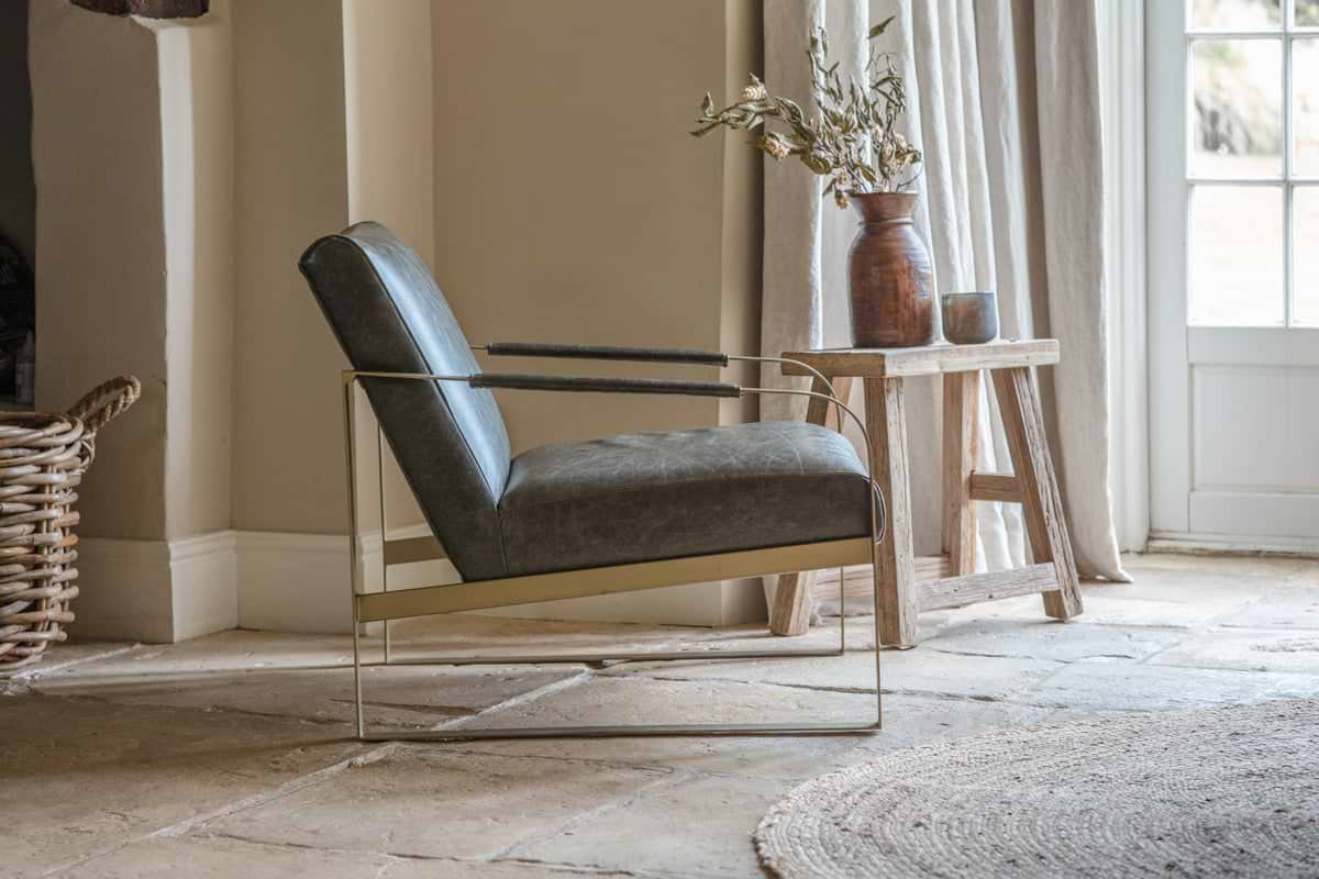 The nkuku Adra Leather & Brass Occasional Chair in Moss Green, featuring slender brass arms and legs, sits on a stone floor by a rustic wood table adorned with a brown vase of dried flowers and a ceramic jug. Sunlight filters through glass panes behind.