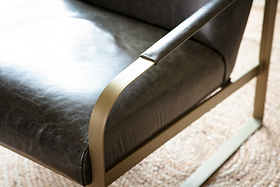 Close-up of the nkuku Adra Leather & Brass Occasional Chair in moss green, featuring a smooth leather seat and backrest with sleek curved antique brass armrests and frame, placed on a neutral woven rug.