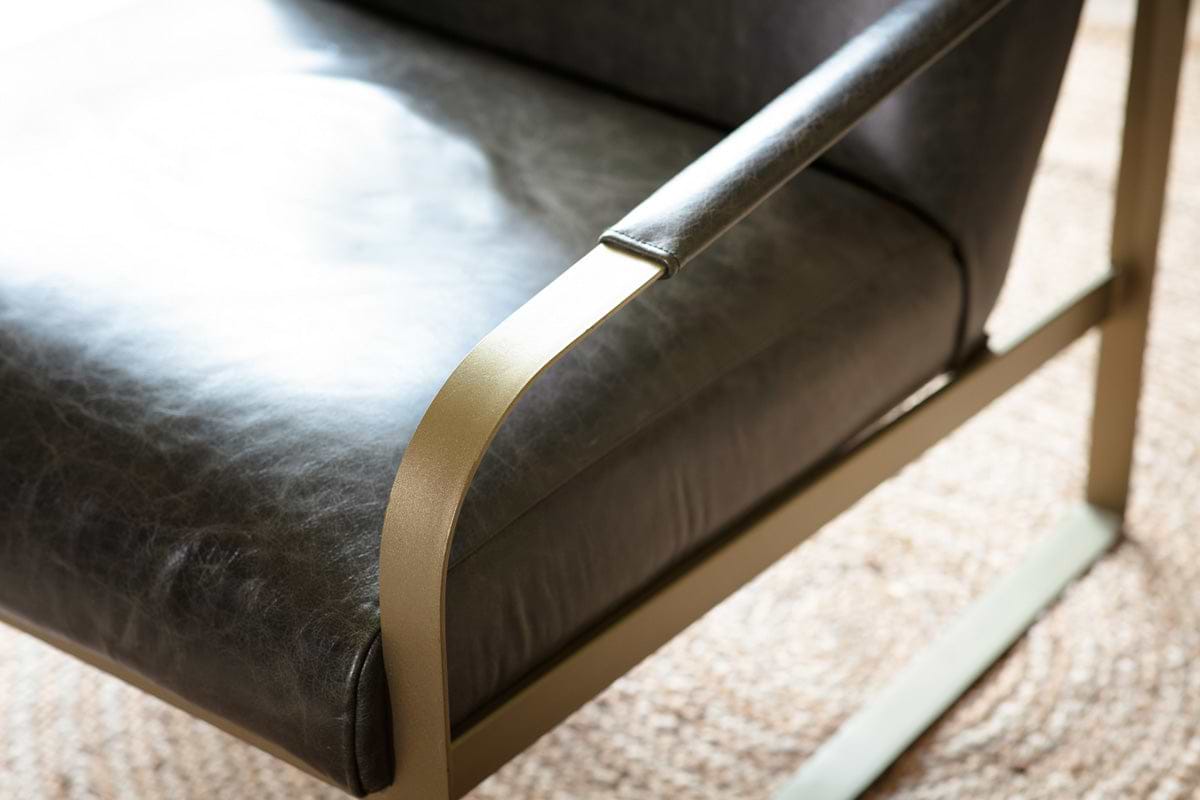 Close-up of the nkuku Adra Leather & Brass Occasional Chair in moss green, featuring a smooth leather seat and backrest with sleek curved antique brass armrests and frame, placed on a neutral woven rug.