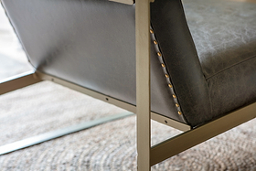 Close-up of the nkuku Adra Leather & Brass Occasional Chair in moss green, featuring buffalo leather upholstery, a matte gold metal frame, and antique brass nailhead trim. The chair sits on a neutral-toned woven rug.