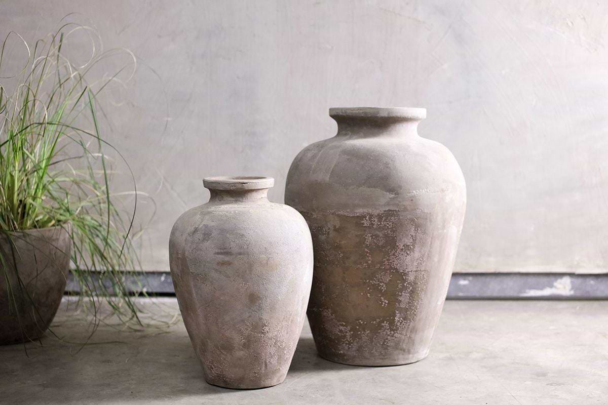 The Affiti Clay Tapered Pot - Small by nkuku sits on a gray concrete floor near a textured pale wall, complemented by a potted plant with slender green leaves for a rustic, minimalist look.