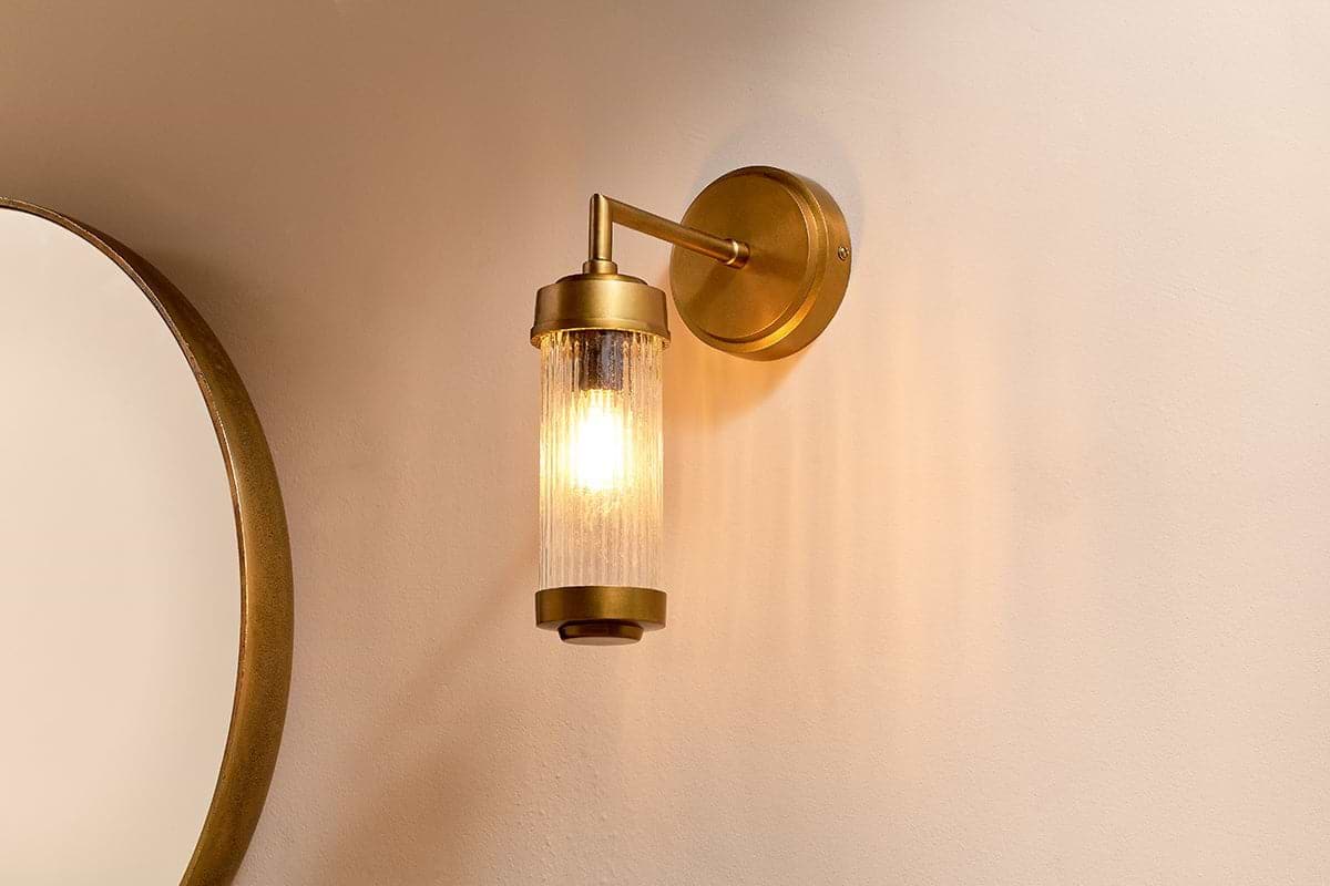 The Akurdi Bathroom Single Wall Lamp by nkuku, featuring an aged brass finish, a round backplate, and a cylindrical glass shade, emits a warm glow against a beige wall. On the left edge of the image, part of a round mirror with a gold rim is visible.