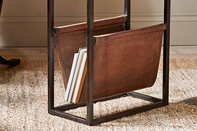 The Alera Side Table by nkuku features a brown leather magazine rack with a black iron frame, holding books and magazines. It sits on a woven beige rug in a room with light walls and wooden floors.