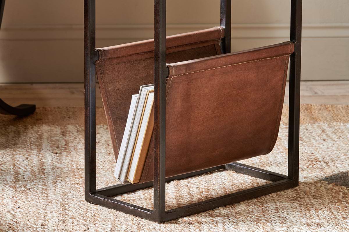 The Alera Side Table by nkuku features a brown leather magazine rack with a black iron frame, holding books and magazines. It sits on a woven beige rug in a room with light walls and wooden floors.