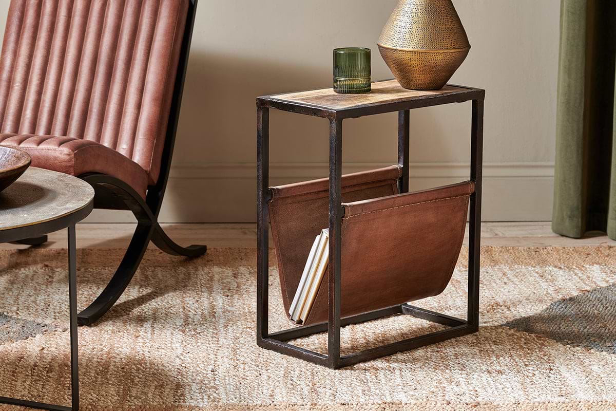 The nkuku Alera Side Table, crafted from rustic mango wood with a leather magazine holder, features a glass and textured bronze vase on top and is placed beside a brown leather chair.
