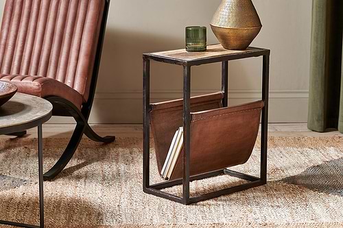 The nkuku Alera Side Table, crafted from rustic mango wood with a leather magazine holder, features a glass and textured bronze vase on top and is placed beside a brown leather chair.
