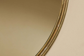 Close-up of the Almora Round Mirror - Large by nkuku, showing its gold-rimmed edge against a beige wall. The mirror’s curved frame and its soft reflection blend with the background, with only part of the frame visible on the right.