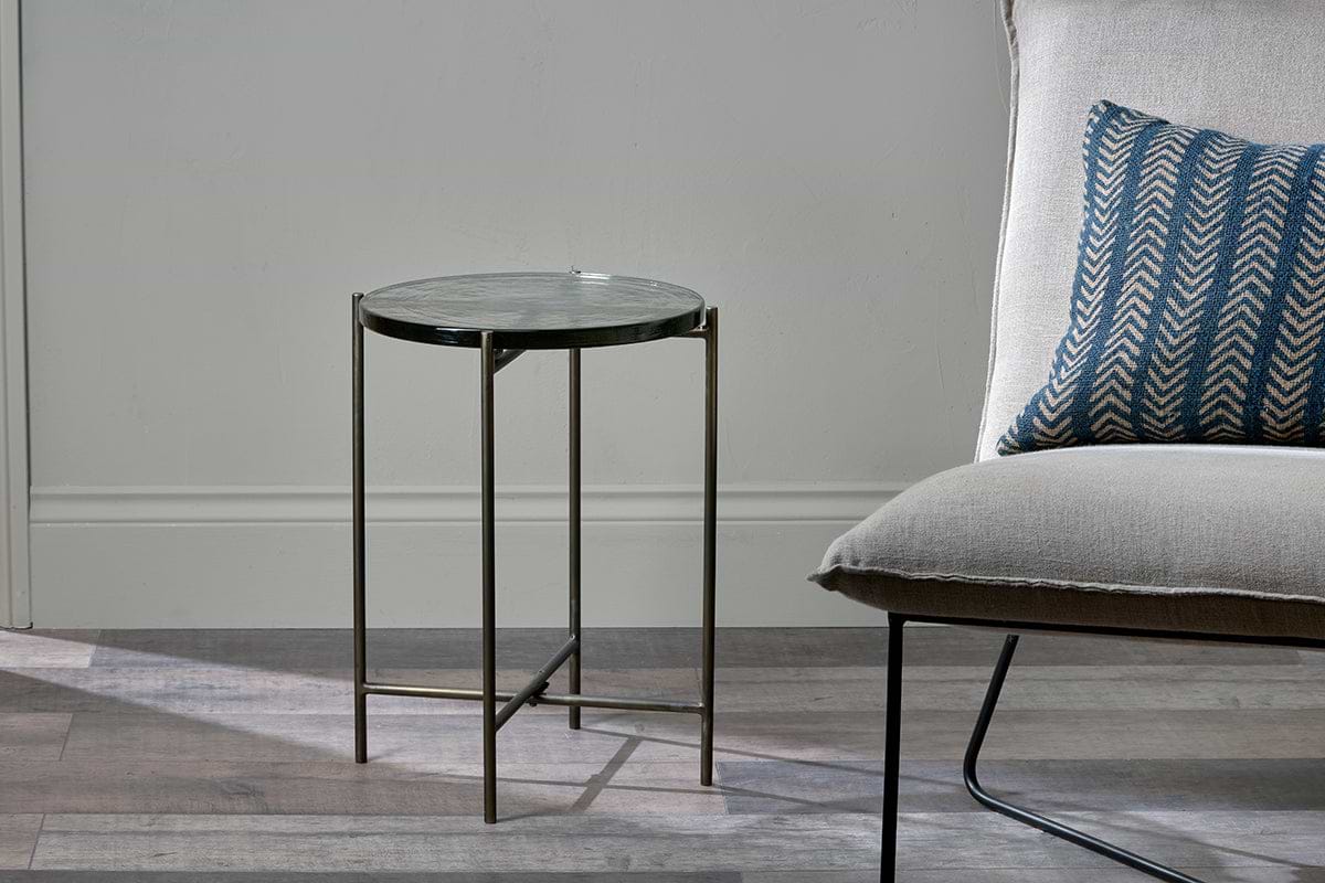 The nkuku Aluva Glass Side Table in clear glass and antique brass sits on a grey wood floor beside a cream wingback chair with a blue chevron cushion, set against a pale grey wall.