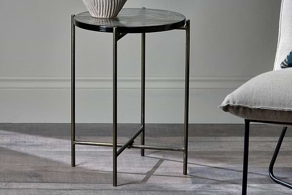 The nkuku Aluva Glass Side Table in clear and antique brass, featuring thin metallic legs, stands on a wooden floor beside an upholstered chair, topped with a ribbed ceramic vase and bathed in natural light from the left.