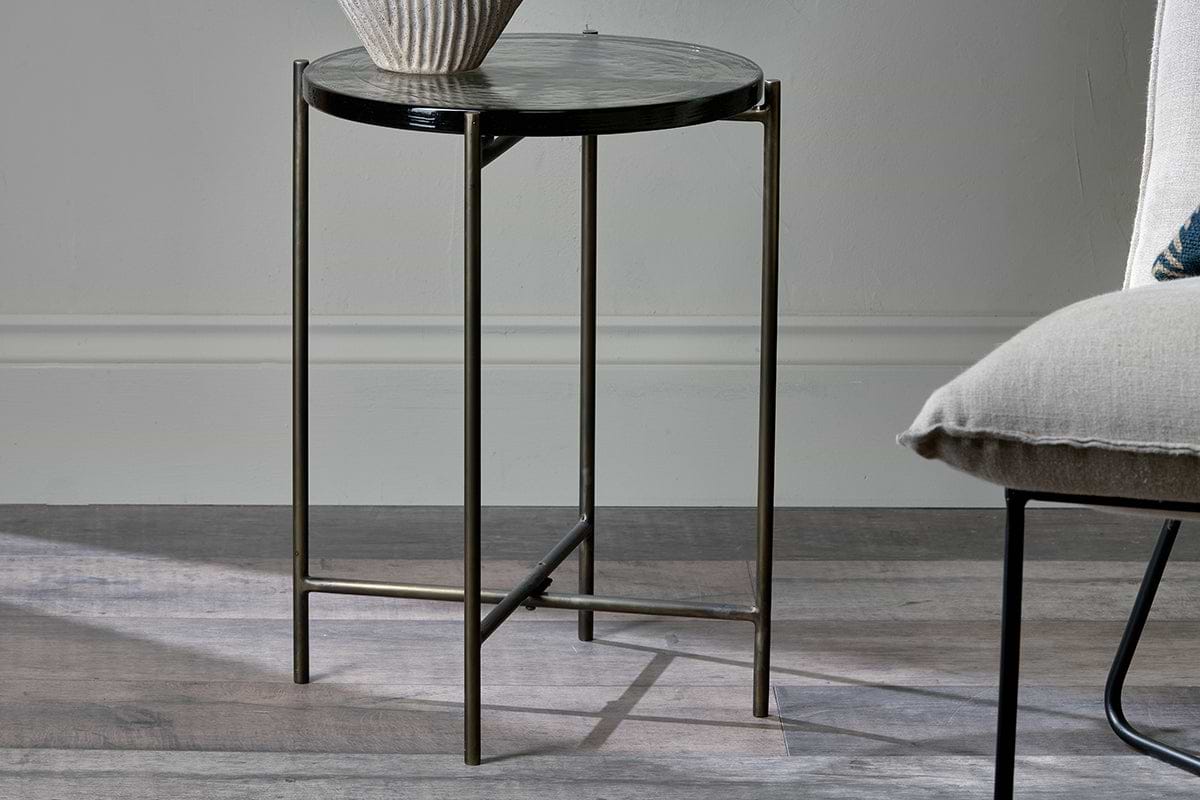 The nkuku Aluva Glass Side Table in clear and antique brass, featuring thin metallic legs, stands on a wooden floor beside an upholstered chair, topped with a ribbed ceramic vase and bathed in natural light from the left.