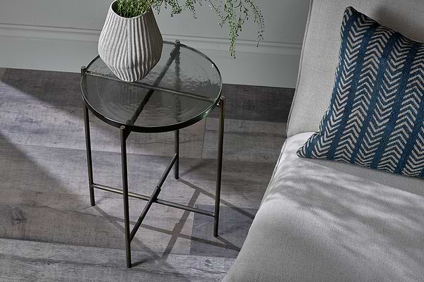 The nkuku Aluva Glass Side Table in clear and antique brass stands beside a light sofa, topped with a striped ceramic vase of green foliage. Sunlight highlights the sand cast glass and a blue chevron cushion on the wooden floor.