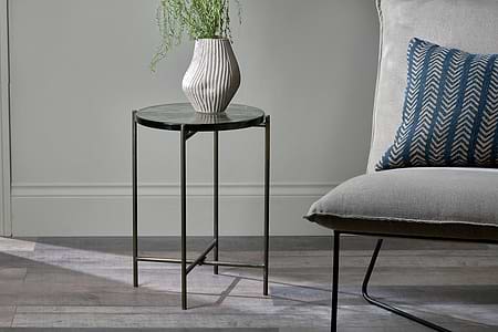 The nkuku Aluva Glass Side Table in clear and antique brass holds a ribbed beige vase with green foliage. Beside it sits a chair featuring a light gray linen cushion and a navy blue chevron throw pillow.