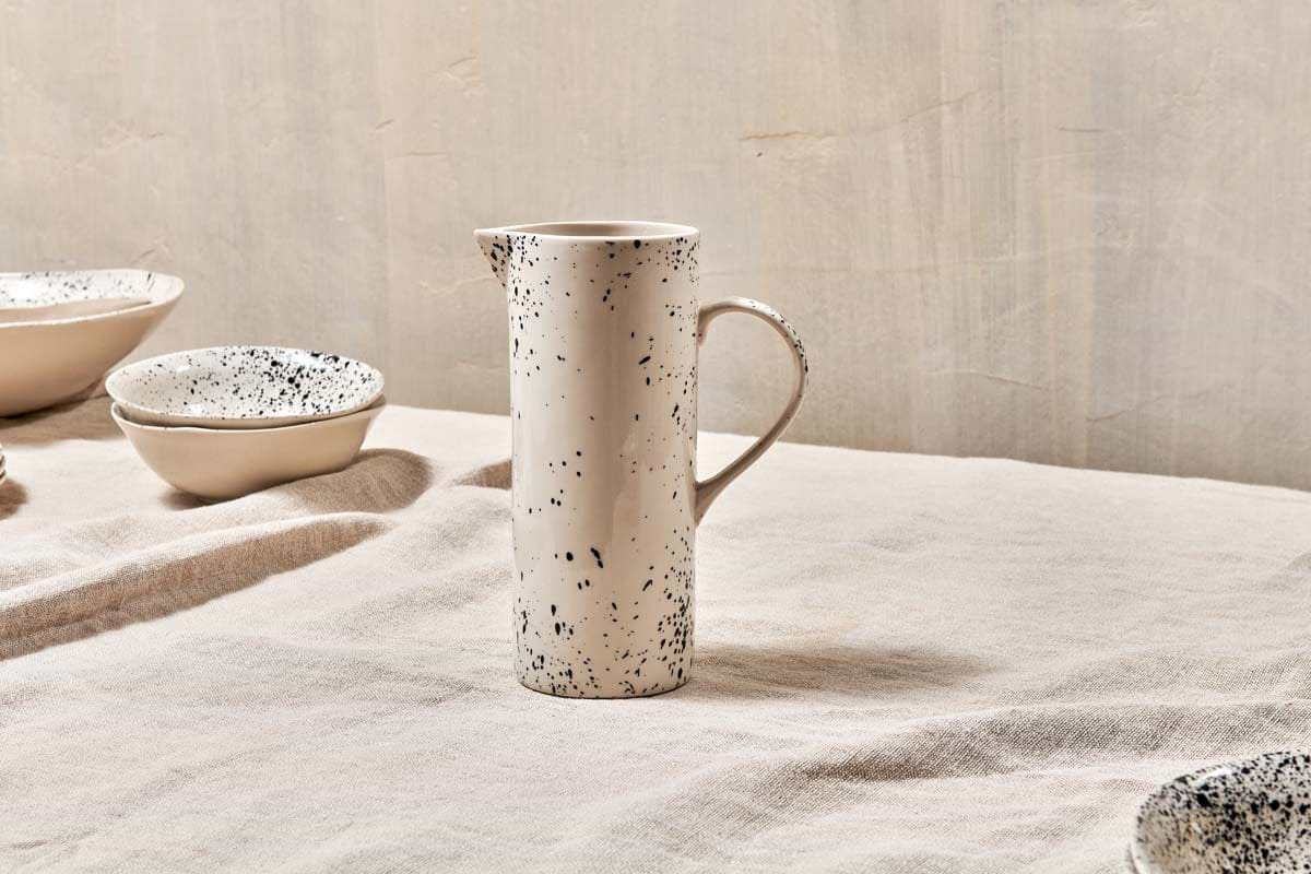 The nkuku Ama Splatter Jug, a tall white ceramic pitcher with a black splatter pattern and curved handle, sits on a beige textured tablecloth. Matching speckled bowls are partially seen against a neutral wall in the background.