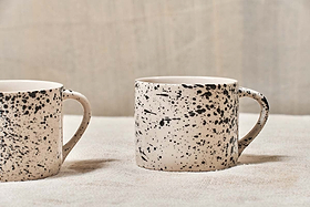 A set of two nkuku Ama Short Splatter Mugs with handmade black splatter designs sit on a light textured surface. Only part of the left mug is seen, the right mug is fully visible, both handles facing right against a neutral background.