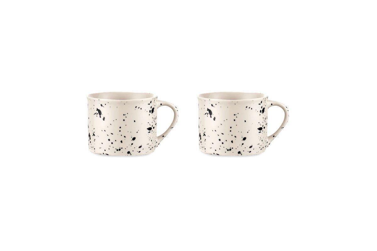 The Ama Short Splatter Mug Set by nkuku features two white handmade mugs with black splatters, rounded bodies, and large handles, displayed side by side on a plain white background in a minimalist, well-lit photo.