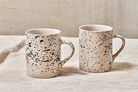 Two nkuku Ama Mug - Tall ceramics with a black splatter finish stand side by side on textured, light fabric. The softly blurred, neutral background highlights the artistic, handcrafted design of these unique mugs.