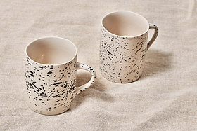 Two cream-colored nkuku Ama Mug - Tall mugs with bold black splatter finish rest on textured beige fabric. Both handmade mugs feature handles and a similar design, with the left one placed slightly closer to the camera than the right.