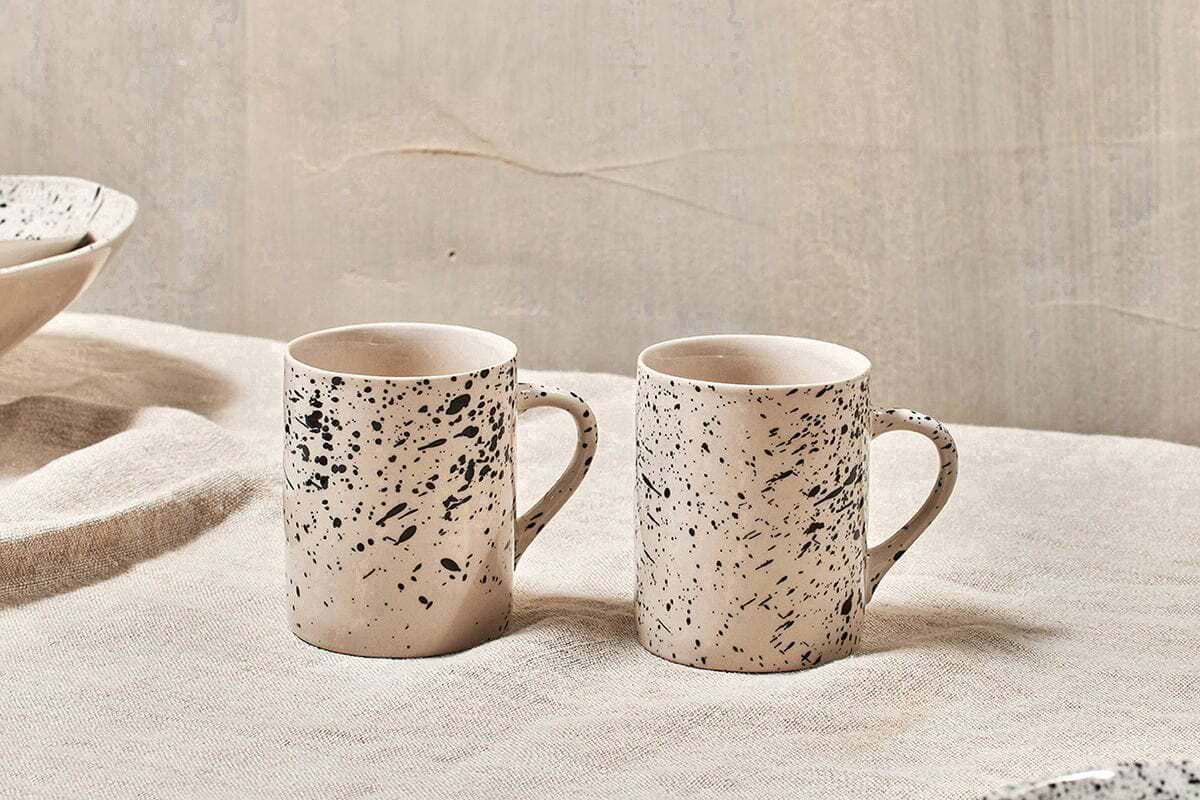 Two nkuku Ama Mug - Tall pieces in off-white with a black splatter finish sit side by side on a beige textured fabric, the neutral backdrop enhancing the minimalist and rustic look of these artisan mugs.