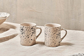 Two nkuku Ama Mug - Tall pieces in off-white with a black splatter finish sit side by side on a beige textured fabric, the neutral backdrop enhancing the minimalist and rustic look of these artisan mugs.
