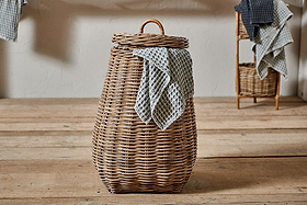 The Amandi Rattan Laundry Bin by nkuku stands on a wooden floor with a blue and white checkered cloth draped over its side. In the background are stacked towels and a smaller natural rattan basket against a plain wall.