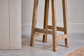 A close-up of the Ambi Wooden Stool - Tall by nkuku shows its handcrafted mango wood legs and natural grain on a smooth gray floor, with part of a textured cream table and woven rug nearby against a plain wall with molding.