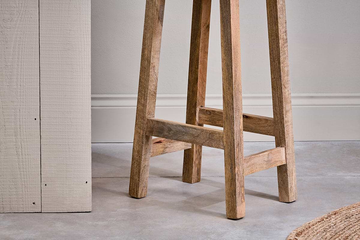 A close-up of the Ambi Wooden Stool - Tall by nkuku shows its handcrafted mango wood legs and natural grain on a smooth gray floor, with part of a textured cream table and woven rug nearby against a plain wall with molding.