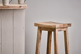 The nkuku Ambi Wooden Stool - Tall, featuring a curved mango wood seat and sturdy legs, stands beside a rustic light wood counter. Two handled ceramic mugs sit on the counter against a plain light gray wall.
