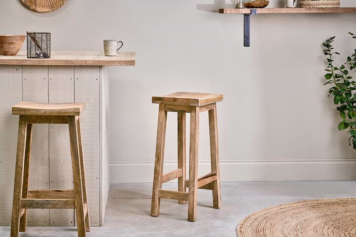 Two Ambi Wooden Stool - Tall by nkuku with square seats stand by a pale wood kitchen island. A cup and decor sit on the counter, with a round woven rug, leafy green plant, and muted accents on wooden shelves behind.