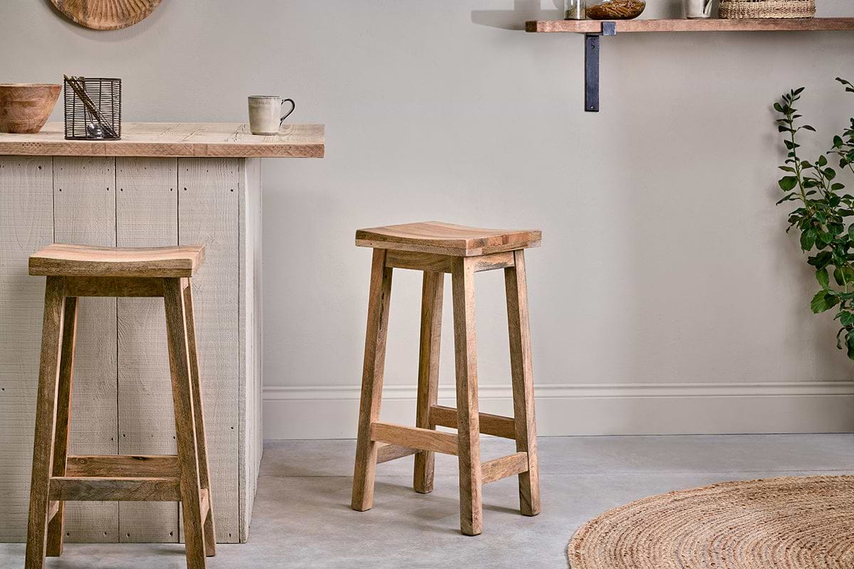 A minimalist kitchen featuring two nkuku Ambi Wooden Stools - Tall by a light wood counter. A mug, decor, woven rug, green plant, and a floating shelf with jars and bowls add warmth to the neutral space.