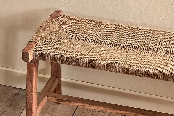The Amhara Acacia & Jute Woven Bench by nkuku features a jute seat and simple acacia wood frame with rectangular legs, displayed against a cream wall on a wooden floor, highlighting its rustic, minimalist design and natural materials.