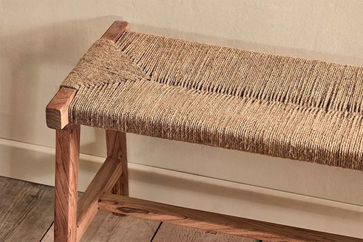 The Amhara Acacia & Jute Woven Bench by nkuku features a jute seat and simple acacia wood frame with rectangular legs, displayed against a cream wall on a wooden floor, highlighting its rustic, minimalist design and natural materials.